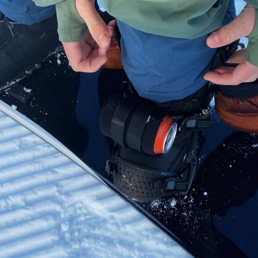 Black beer can holder strapped to a snowboard binding's front strap, holding a 12 oz can.