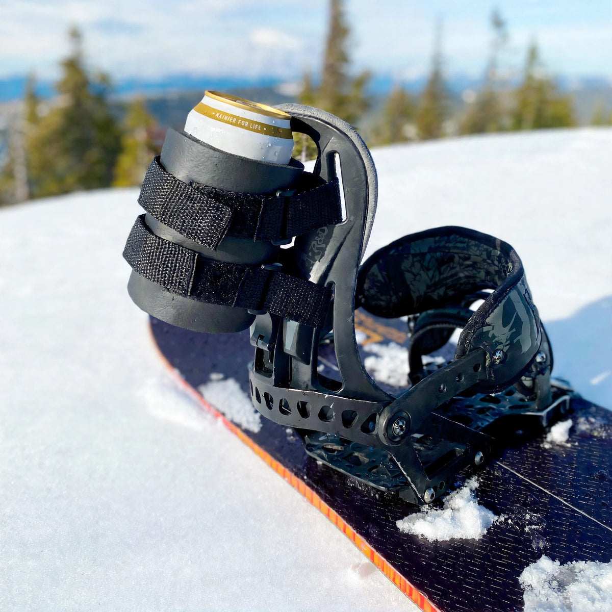 Black beer can holder strapped to a snowboard binding's highback, holding a Rainier beer can with a mountain backdrop.