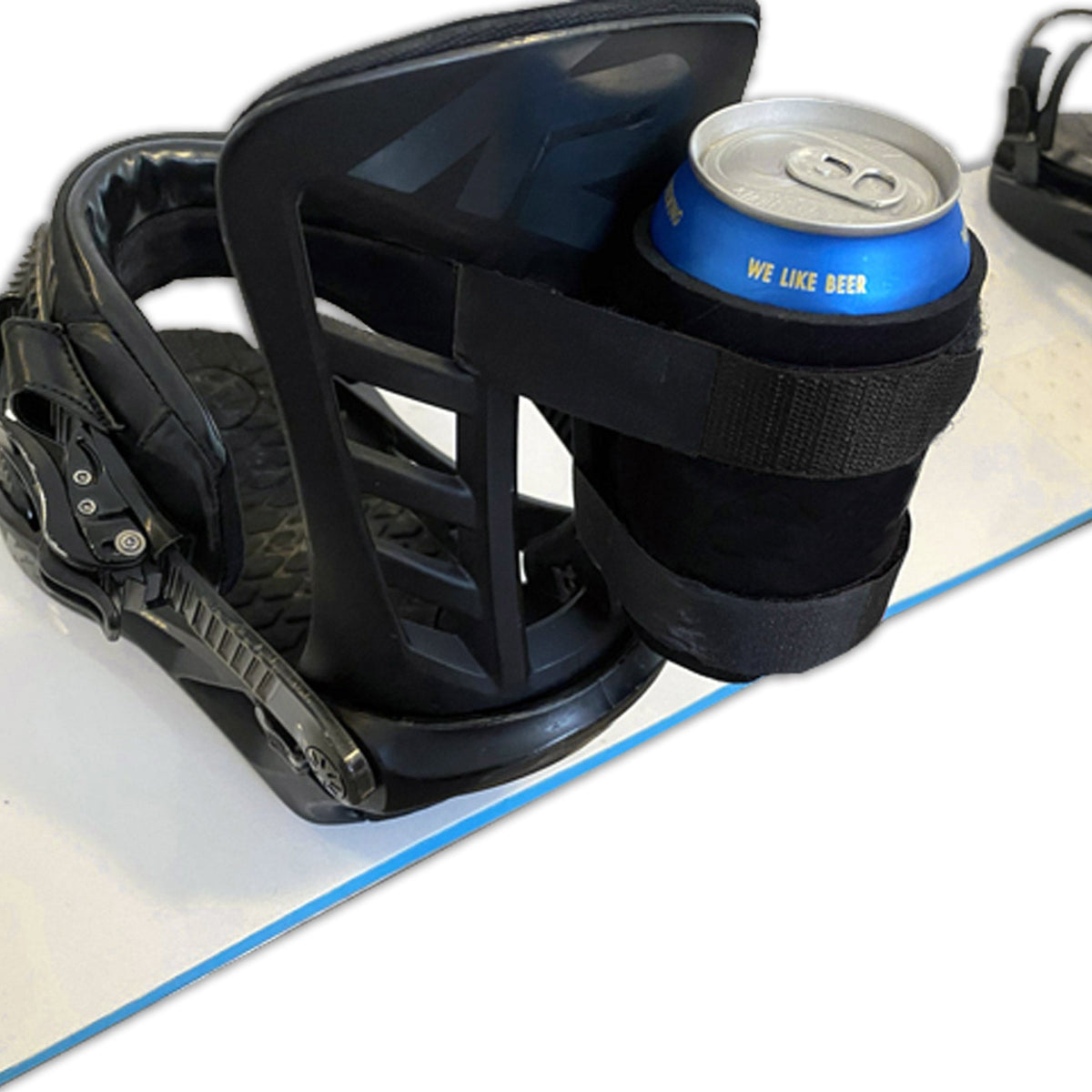 Black beer can holder strapped to a snowboard binding, holding a blue can on a snowboard.