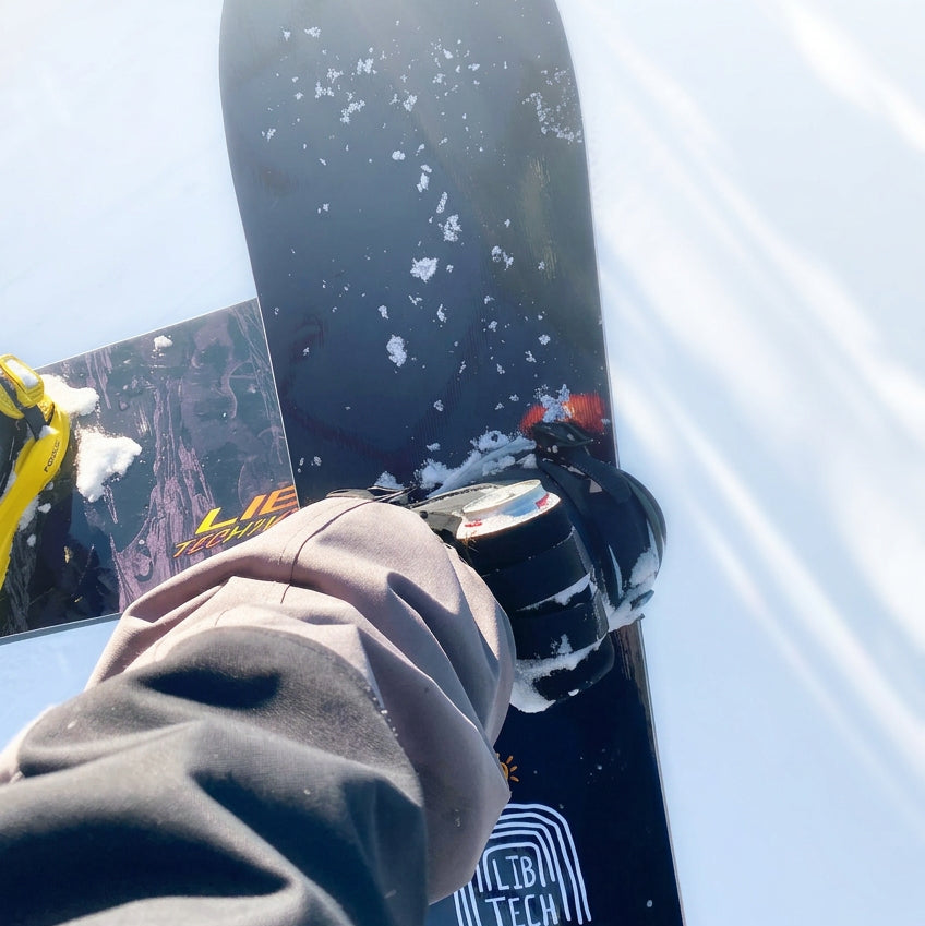 First-person view of a black beer can holder strapped to a snowboard binding, holding a can on the lift.