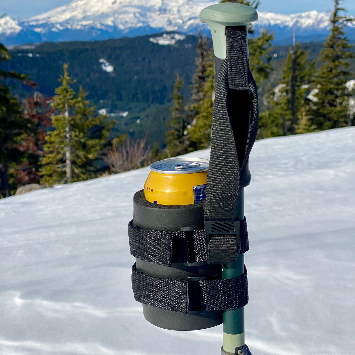 Black beer can holder strapped to a ski pole, holding a yellow can with snowy mountains behind it.