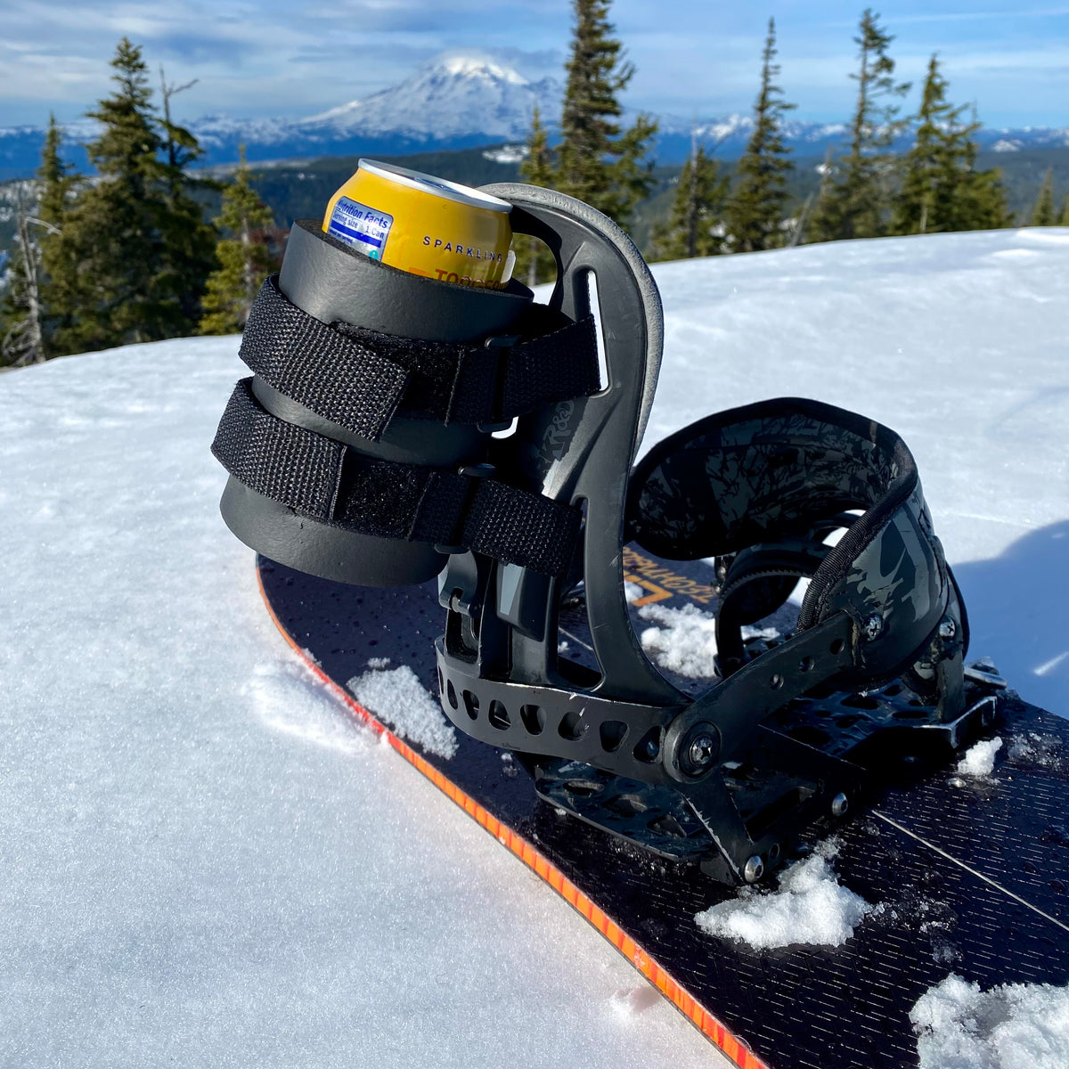 Black beer can holder strapped to a snowboard binding's highback, holding a yellow can with a mountain backdrop.
