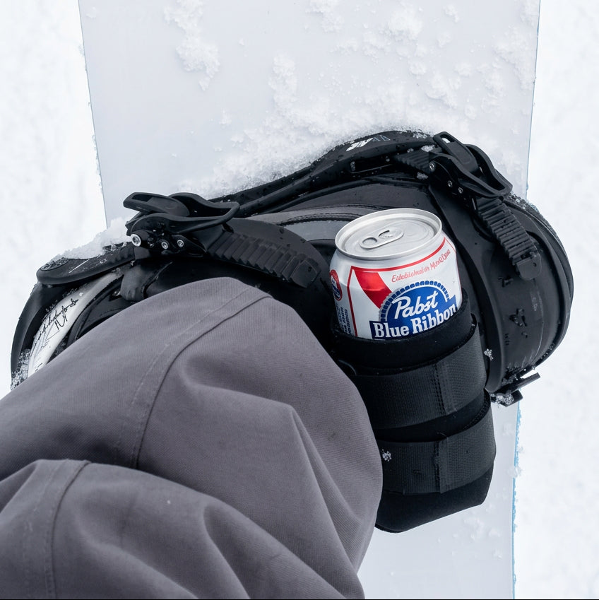 Black beer can holder strapped to a snowboard binding, holding a 12 oz can on the slopes.