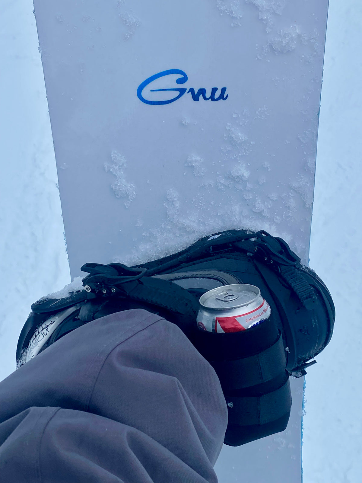 Black beer can holder strapped to a snowboard binding, holding a 12 oz can on the slopes.