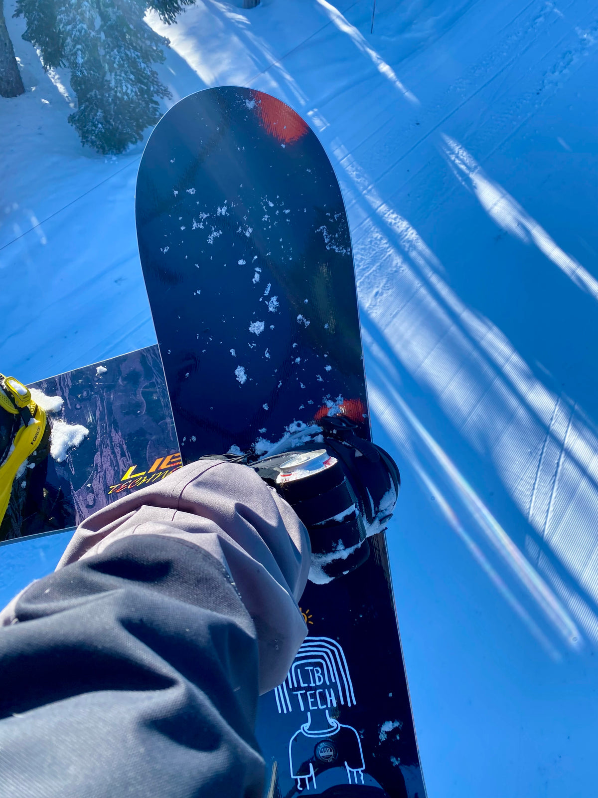 First-person view of a black beer can holder strapped to a snowboard binding, holding a can on the lift.